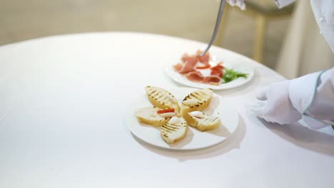 professional cook decorates toasts with slices of meat, tomatoes by kitchen pinchers. beautiful presentation of tasty toasts on a white plate for guests