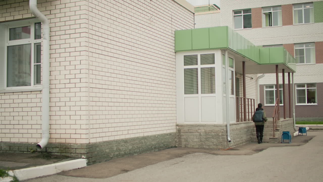 Мальчик с рюкзаком бежит к зданию школы, замедляясь у ступеней, готовясь подняться ко входу, демонстрируя решимость и готовность к учебному дню в городских условиях