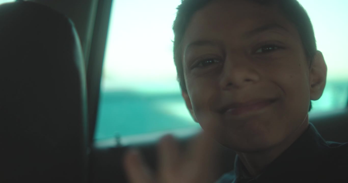 Smiling boy poses inside car with river and boat visible through window behind him