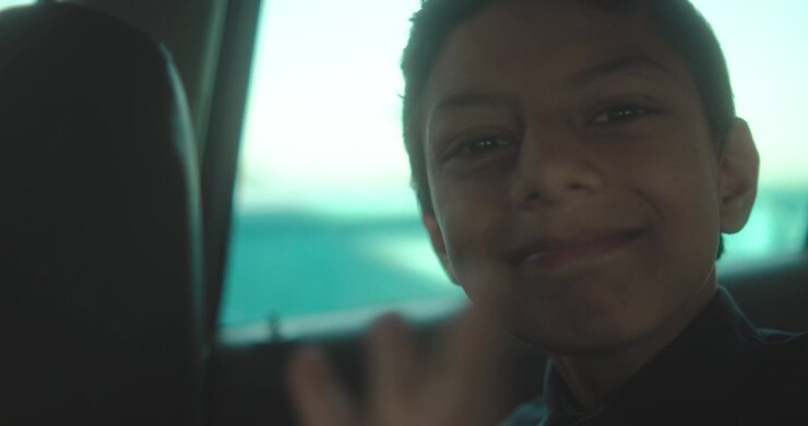 Smiling boy poses inside car with river and boat visible through window behind him
