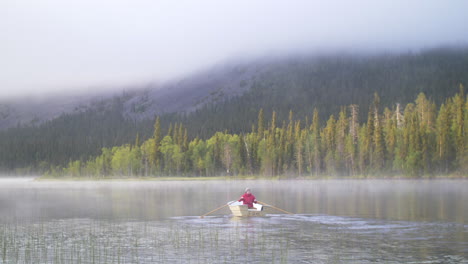 Young-man-rowing-a-small-boat-on-a-misty-lake-with-green-forest-and-mountain-in-the-background