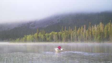 young man rowing a small boat on a misty lake with green forest and mountain in the background