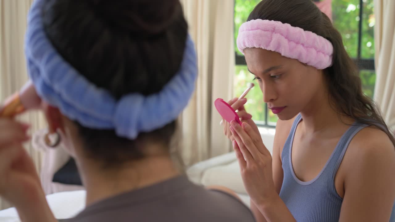 Diverse donne che indossano fasce per capelli, guardano un vestito rosa e si applicano l'ombretto con uno specchietto compatto, a casa.