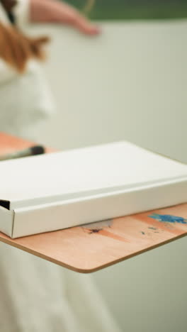 a close-up shot of a person holding a wooden palette with paintbrushes and a white paint box, preparing to paint outdoors. the individual wears a white t-shirt with a green and blue checkered flannel