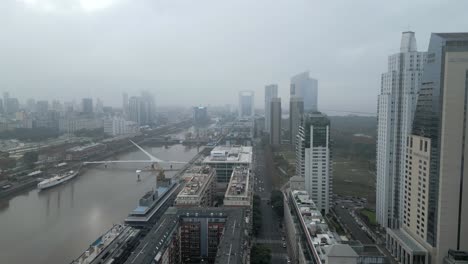 Vista-Aérea-Panorámica-De-Edificios-Altos-A-Lo-Largo-Del-Río-De-La-Plata-Con-El-Puente-De-La-Mujer-Bajo-El-Cielo-Nublado-Debido-Al-Cambio-Climático,-Buenos-Aires,-Argentina