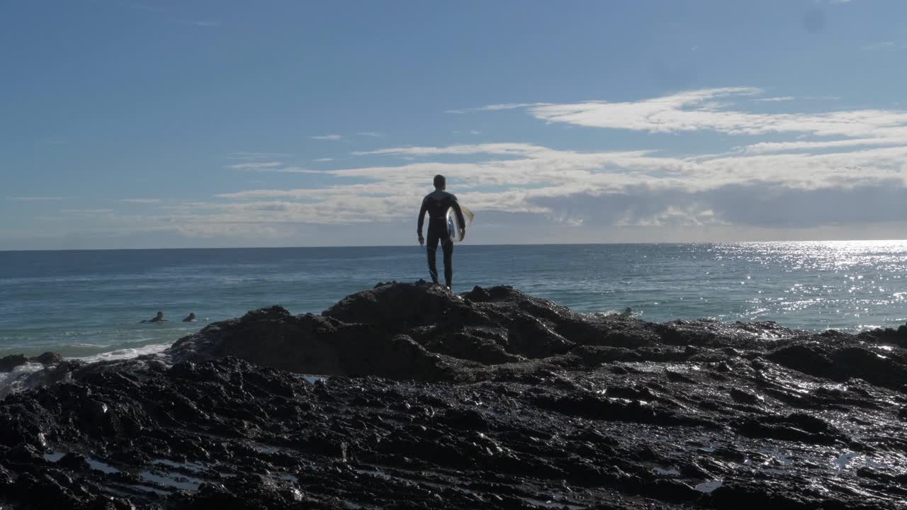 hombre con tabla de surf se encuentra en las rocas con olas salpicadas - surf en rocas de pargo - coolangatta, costa dorada, australia
