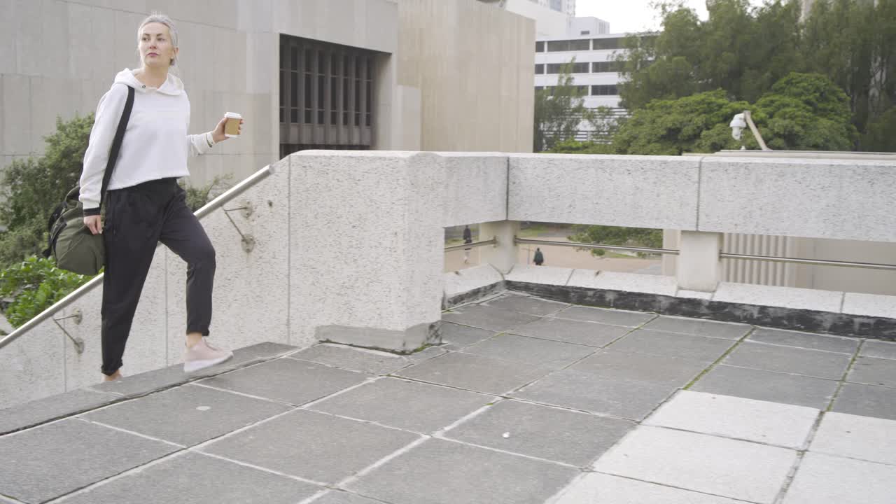 Woman ascending rooftop steps hearing ringtone, answering call carrying coffee, olive duffel bag