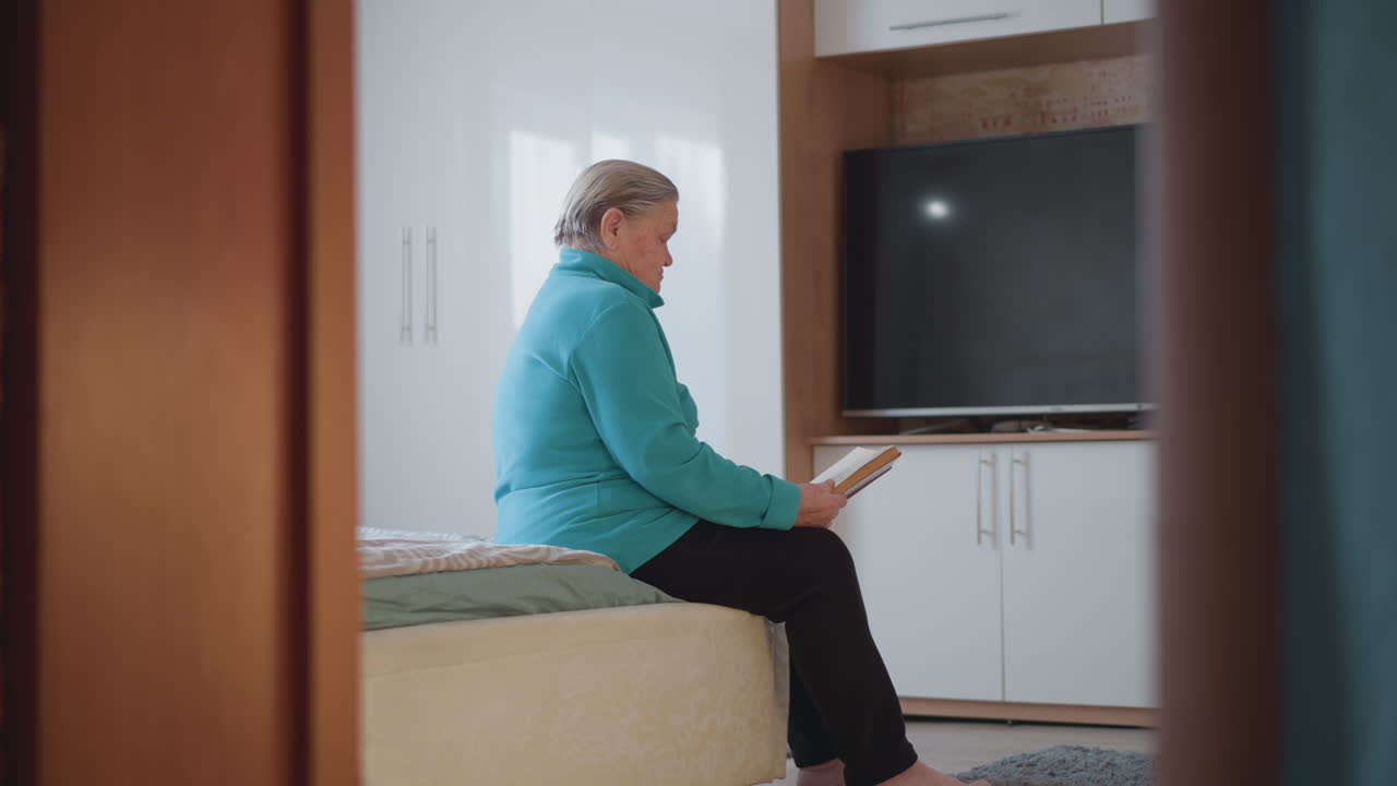 Elderly woman in teal green sweater sitting on bed reading a book, facing sideways, with a TV in the background, peaceful bedroom atmosphere, soft natural light streaming through window, quiet moment