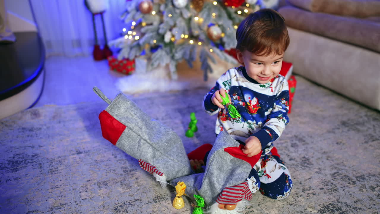 Adorable Caucasian baby sitting on the floor with stockings. Happy kid pulls the sweets and smiles cutely.