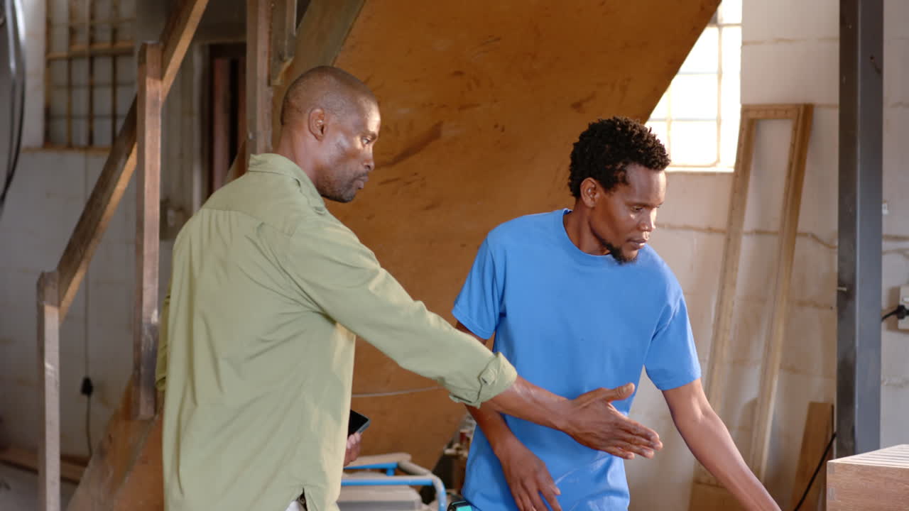 Collaborating African American colleagues reviewing tablet in workshop, inspecting wooden planks