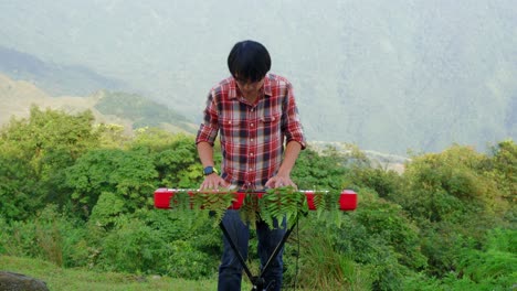 a stationary footage of a man, playing his electronic musical keyboard passionately in the forest near the mountains