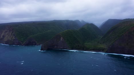 towering green cliffs wrapped in drifting mist loom over restless blue sea, creating a wild and secluded coastal panorama under soft gray light.