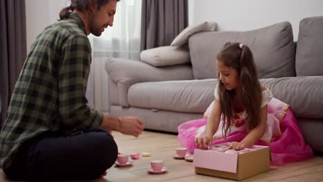 A-little-girl-in-a-fluffy-pink-dress-together-with-her-brunette-male-father-in-a-green-checkered-shirt-lay-out-toy-cups-for-tea-drinking-while-playing-at-home-in-a-modern-apartment