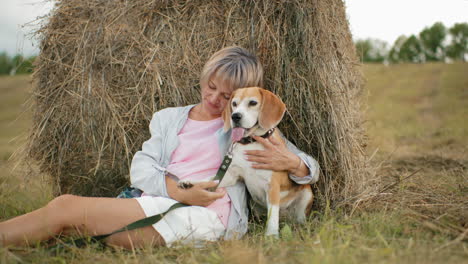 woman holds her dog closely, touching it affectionately while resting against hay bale in vast farmland, a peaceful, loving moment surrounded by open fields