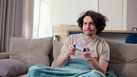 Happy-brunette-guy-with-curly-hair-and-mustache-plays-video-games-using-a-white-joystick-while-sitting-on-a-sofa-with-a-blue-blanket-in-a-modern-apartment