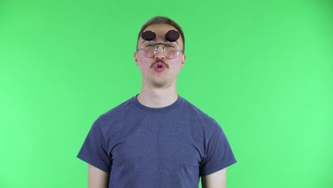 retrato de un joven que abre sus gafas de sol con una expresión entusiasta y sorprendida. un hombre lindo con bigote en una camiseta azul posando en una pantalla verde en el estudio.