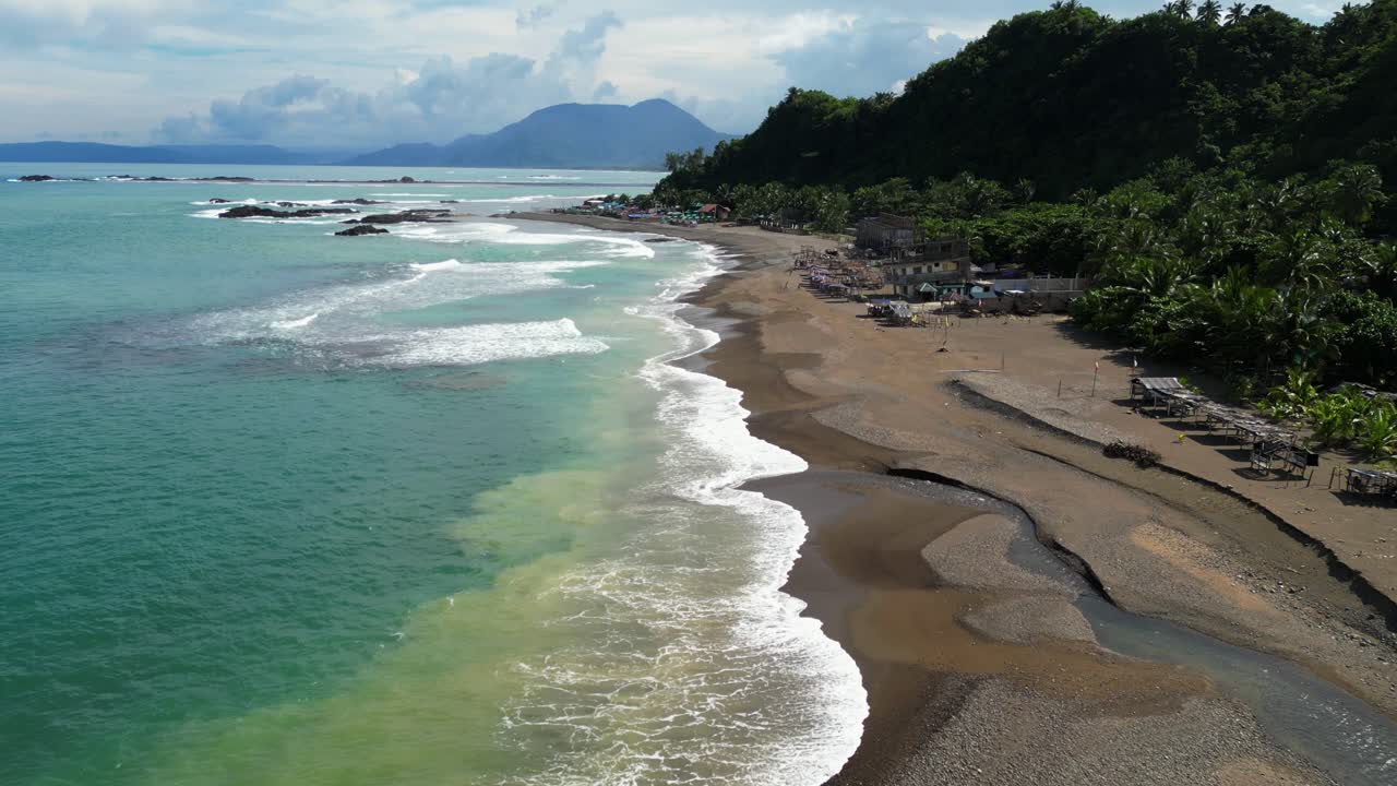 Forward aerial pan in Matawe, Dingalan, Aurora, focusing on waves crashing along the shoreline as turquoise waters meet sandy stretches framed by lush green hills and coastal huts
