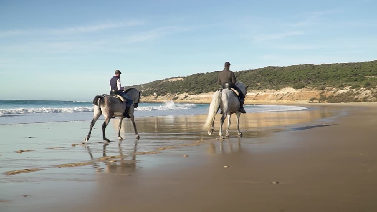 toma panorámica amplia de dos jinetes con sus caballos cabalgando a lo largo de una hermosa playa durante las tranquilas olas del mar