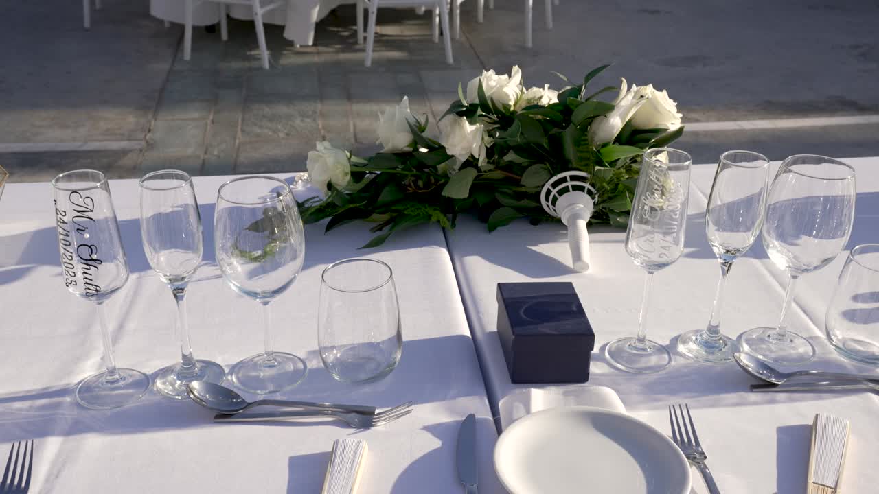 Hvid dug med glas og bestik til en bryllupsceremoni udendørs. Friske blomster er arrangeret midt på bordet.