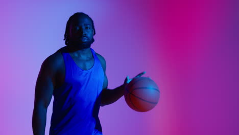 studio portrait of male basketball player wearing team vest bouncing ball against low key colourful mixed lighting