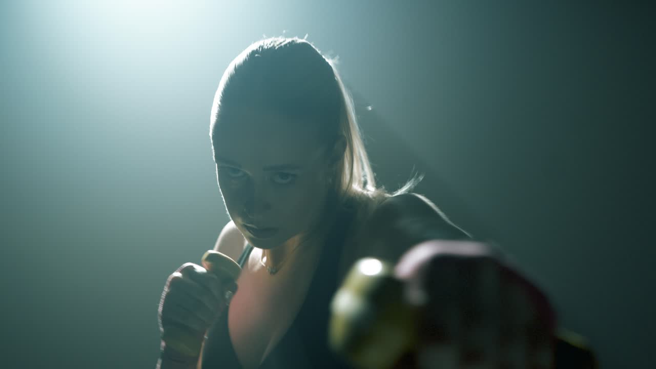 Blonde fit woman shadow boxes and dodges while holding yellow dumbbells. Waist-up tracking shot as she moves toward camera. Backlight creates rim glow in smoky, cinematic training environment