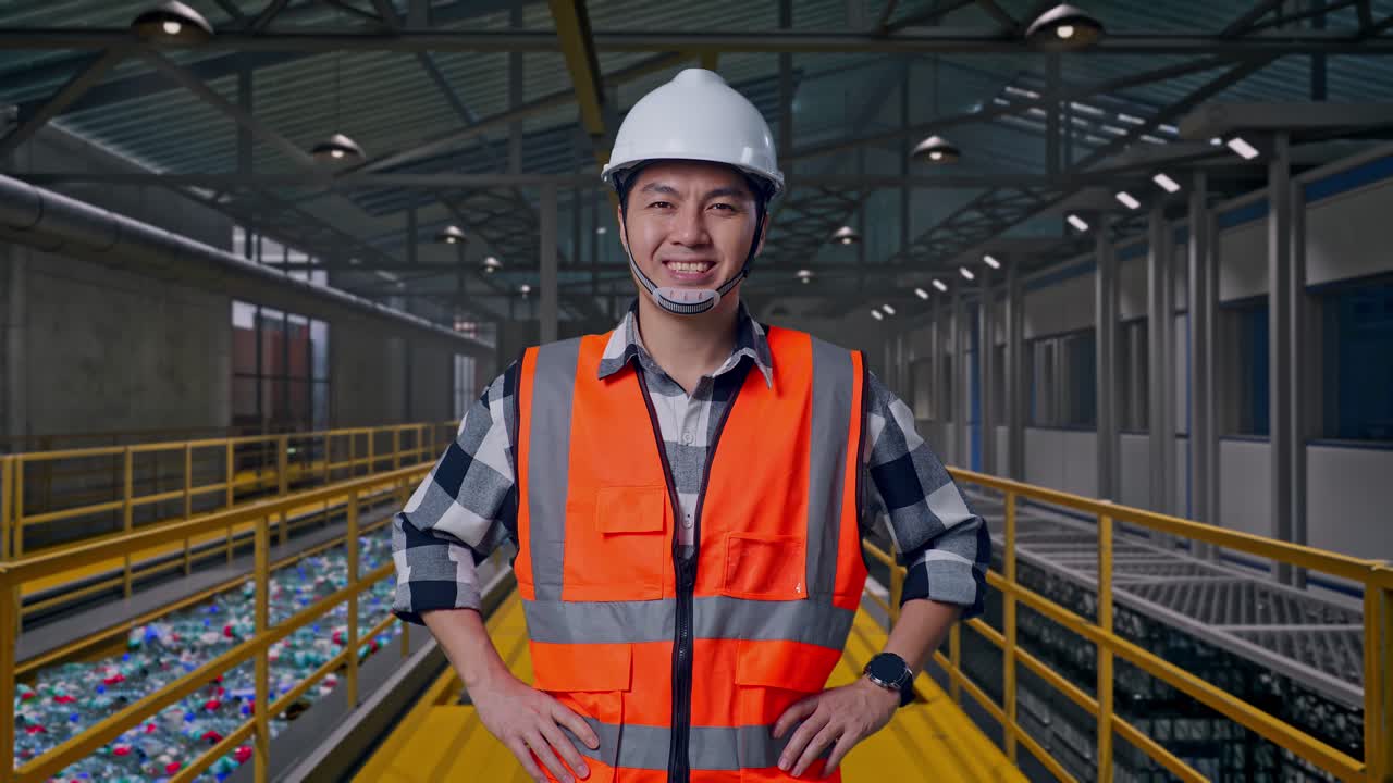 Ingegnere uomo asiatico con casco di sicurezza che sorride alla telecamera mentre è in piedi con le mani sui fianchi, ispezionando le operazioni di un impianto di riciclaggio della plastica con nastro trasportatore di bottiglie.