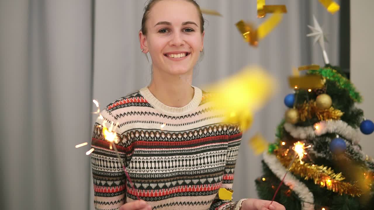 Golden confetti falling on young smiling woman with the Christmas tree and garlands on the background holding sparklers and
