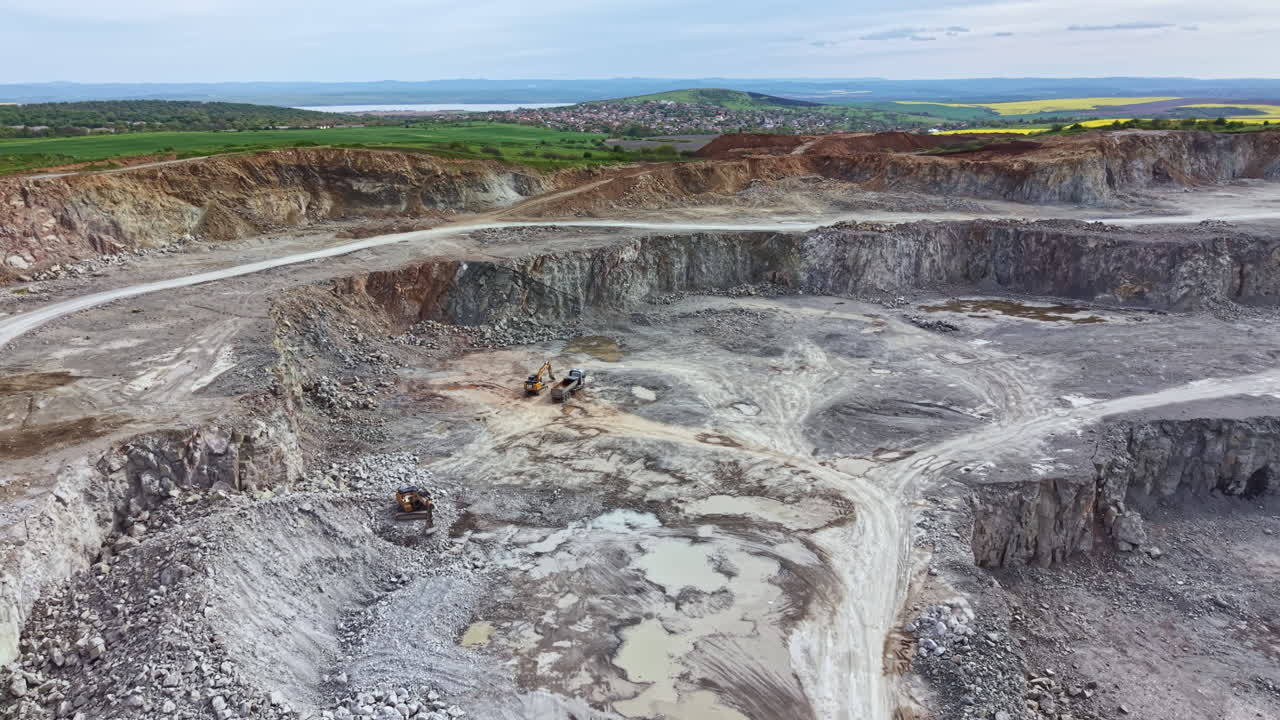 Rock quarry site shows heavy machinery and land being shaped for mining operations near a valley under a cloudy sky