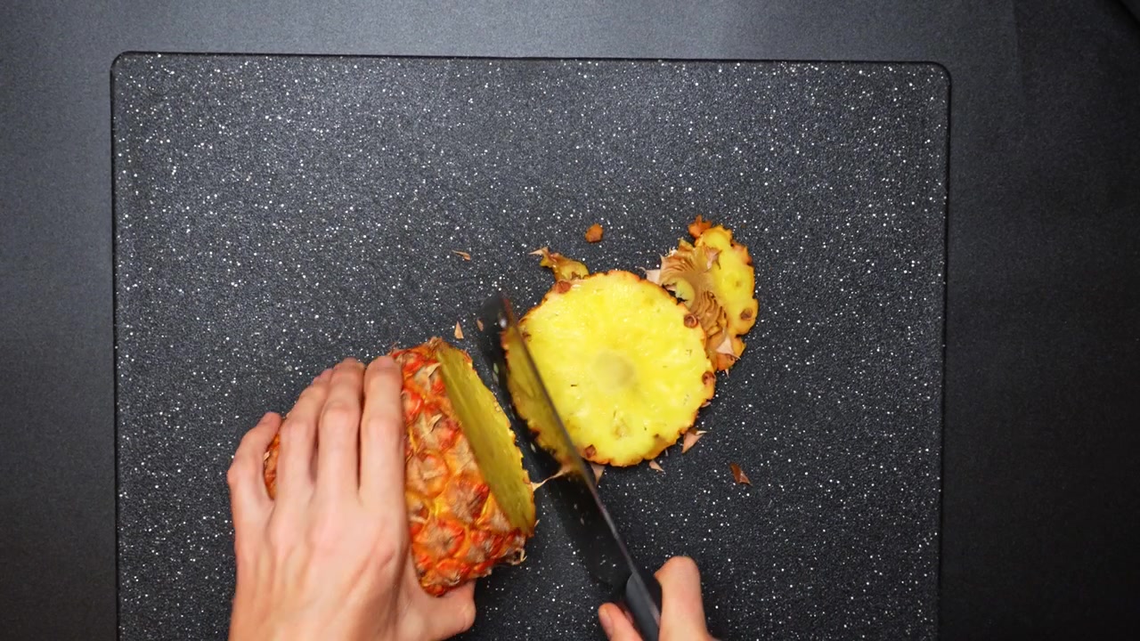 Taglio di un ananas su un tagliere nero in cucina. Delle mani tagliano un ananas fresco su un tagliere nero.