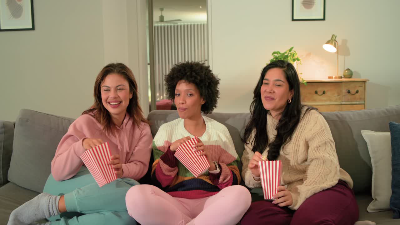 Diverse amiche ridono per un colpo di scena in TV, mangiando popcorn da contenitori a righe sul divano di casa