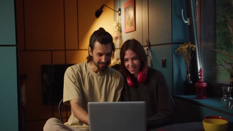 a brunette guy and a brunette girl in red headphones sit and watch a video on a laptop in the evening with a yellow lamp on the background