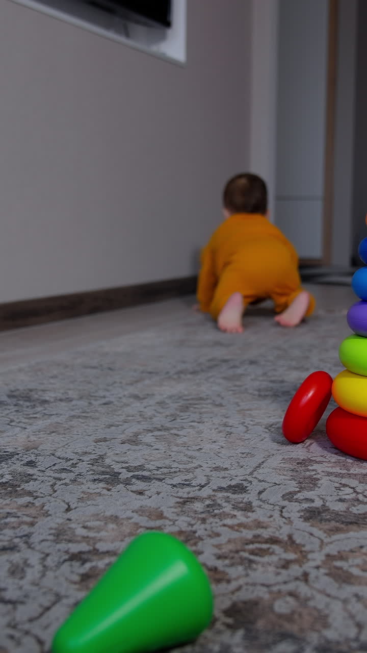 Baby crawling along the room ignoring bright toy pyramid. Boy in orange sport suit stands up holding by the wall. Vertical video