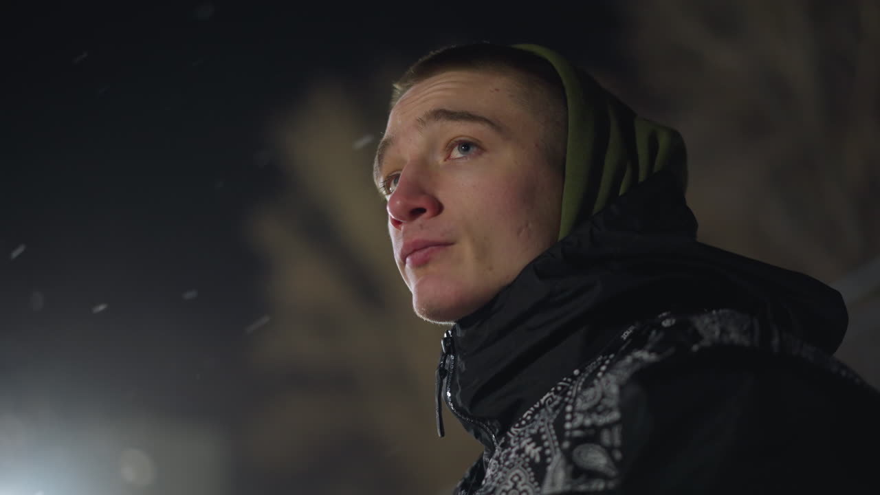 Side profile of student in hoodie standing outdoors during winter night, looking up with visible cold breath, waiting quietly, surrounded by dark sky and softly lit trees in background