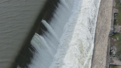 70mm drone footage of a dam spillway with water creating a powerful lateral cascade. the water flows through a wide channel that narrows gradually. the drone flies opposite the water’s path.