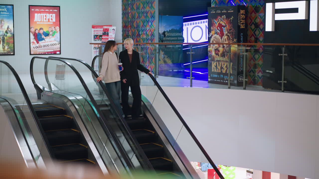 Friends riding escalator inside shopping mall while chatting, one holding drink, both enjoying relaxed moment in bright indoor setting with movie posters and colorful decorations in background
