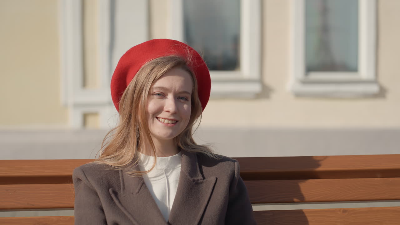 Улыбающаяся женщина на улице. Яркая женщина на городской скамейке. Веселая дама сидит на скамейке в красной шляпе в городской обстановке. Счастливая женщина наслаждается солнечным днем, сидя на деревянной скамейке в красном берете.