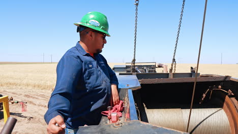 serious hispanic man in blue uniform, glasses and protective helmet works at the oil production site. man holding a lever of equipment unwinding the metal wire.