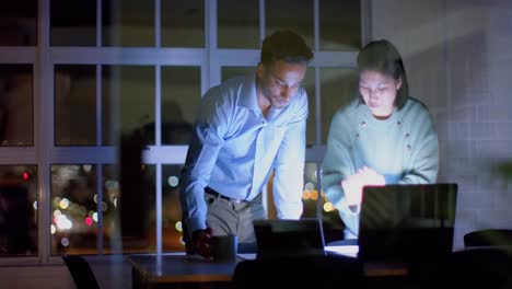 Focused-diverse-male-and-female-colleague-discussing-work-using-tablet-and-laptop-at-night-in-office