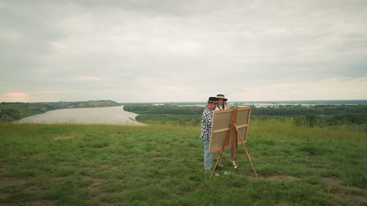 A man in a black hat and plaid shirt stands close to a woman in a white dress, engaged in a discussion or observation in a grassy field with a drawing board set up before them. with a river