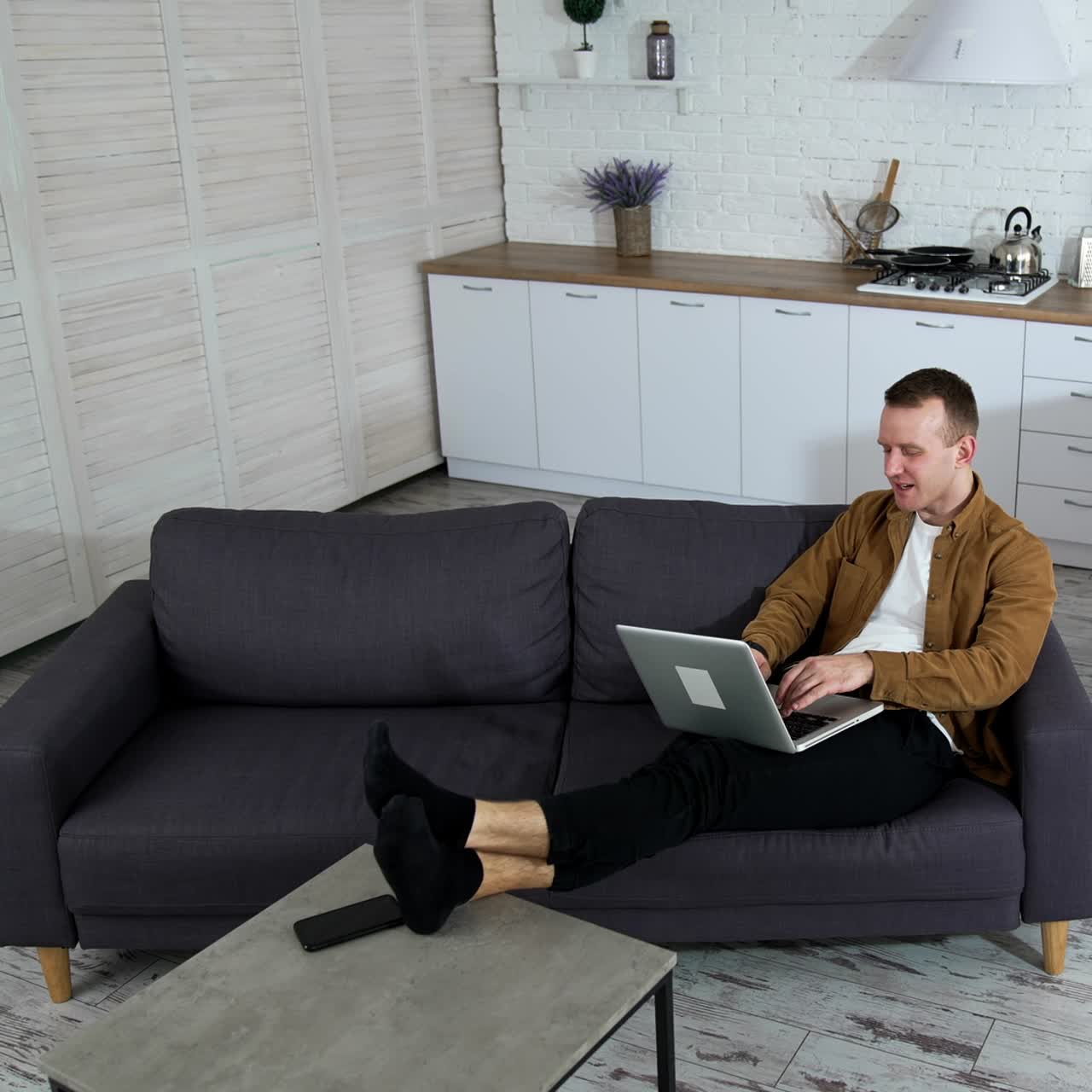 Relaxed guy working on a wireless computer at home. Young man laying on a sofa and typing on a laptop on modern kitchen background. Job at quarantine