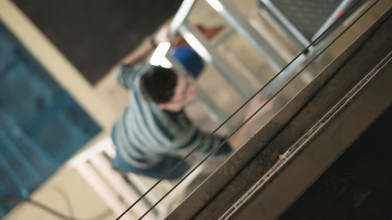 Young technician wearing glasses climbs indoor metal scaffold ladder toward diamond plate platform, overhead perspective shows industrial maintenance action during workshop setup for lighting rig