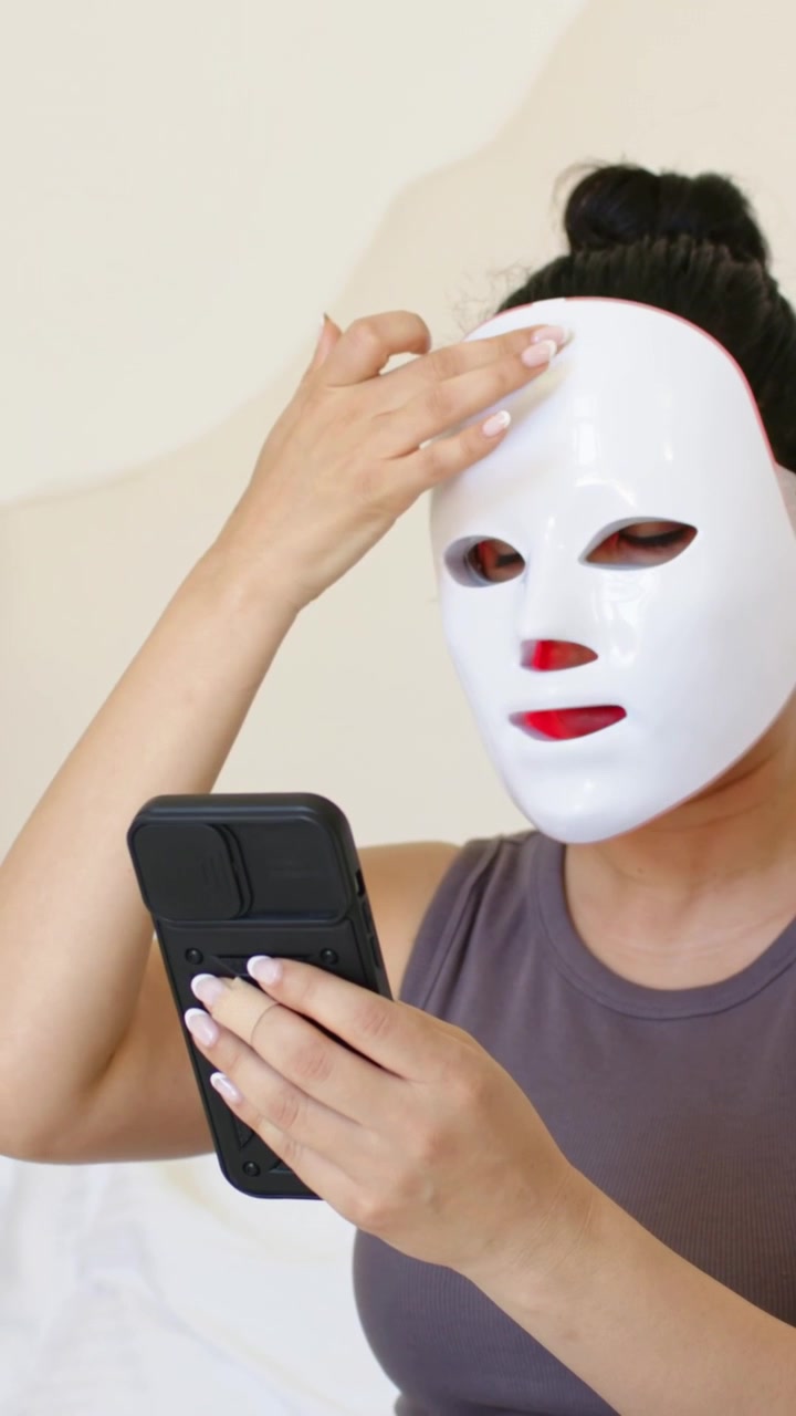 Video vertikal: Wanita mengenakan tank top abu-abu menekan tombol remote sambil menyesuaikan masker wajah di tempat tidur untuk perawatan kulit.