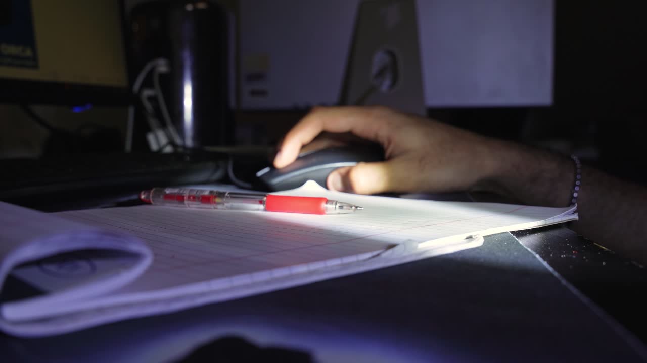 a person writing in a lined notebook with a red pen on a desk under low light, creating a focused indoor study and productivity atmosphere