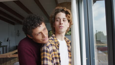 Happy-diverse-gay-male-couple-looking-through-window-and-embracing-at-home,-slow-motion