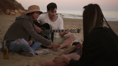 young man is playing guitar by the fire sitting on the beach together with friends