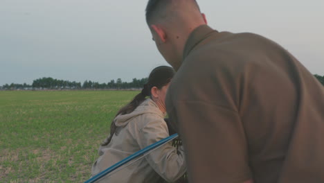 man and woman work together lifting hot air balloon basket during outdoor setup, showing teamwork and preparation against backdrop of green field and soft evening sky