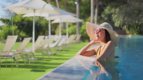 Mujer-En-Traje-De-Baño-En-La-Cubierta-De-La-Piscina-De-Hormigón-Junto-A-La-Piscina-Con-Sillas-Y-Sombrillas-En-Segundo-Plano.