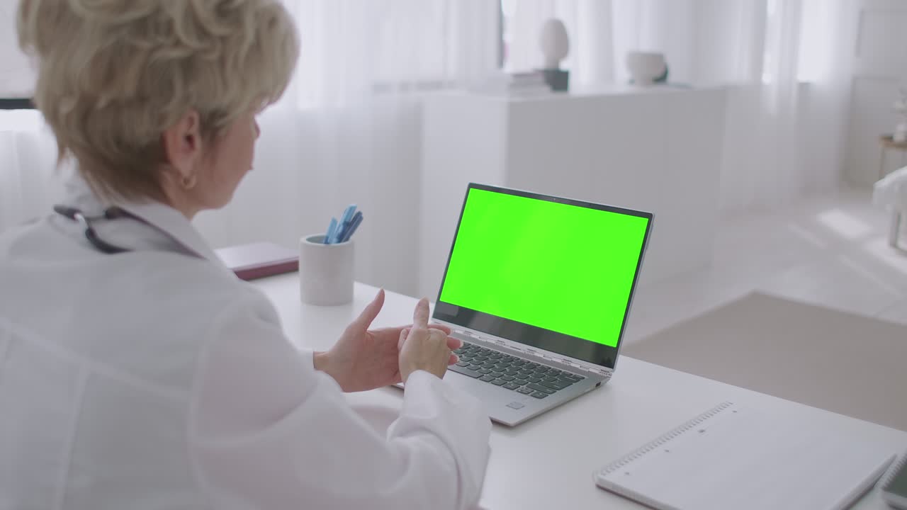 la mujer médico está escuchando al paciente durante la consulta en línea, mirando la pantalla verde de la computadora portátil para la tecnología de llave cromática, trabajando remotamente