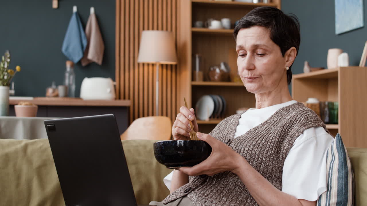 Donna che mangia da una ciotola mentre usa il laptop a casa