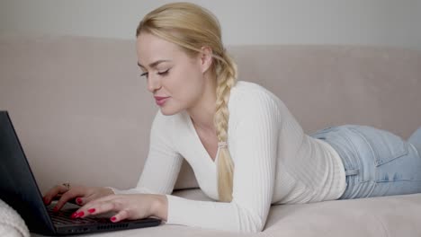 Blond-female-lying-on-sofa-and-browsing-laptop